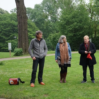 Drei Männer und zwei Frauen stehen in einem Park, rechts ist eine Stele zu sehen, gebildet aus dem Wort „Espero“, die Buchstaben stehen untereinander.