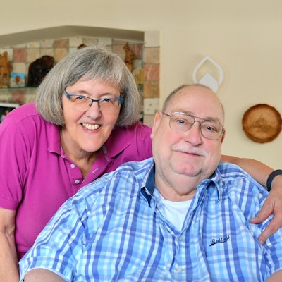 Die Eheleute schauen freundlich in die Kamera. Karin Benden umarmt ihren Mann, der auf einem Sessel sitzt, während sie neben ihm steht.