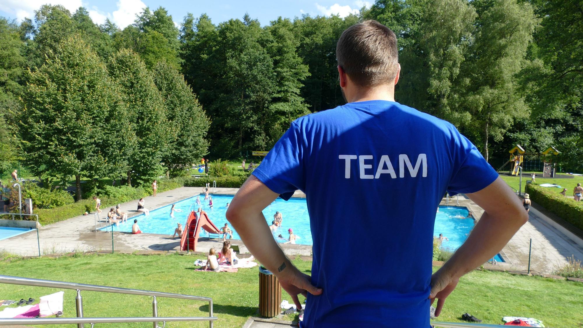 Ein Mann schaut auf ein Schwimmbad. Er trägt ein blaues T-Shirt, auf dem Rücken steht Team.