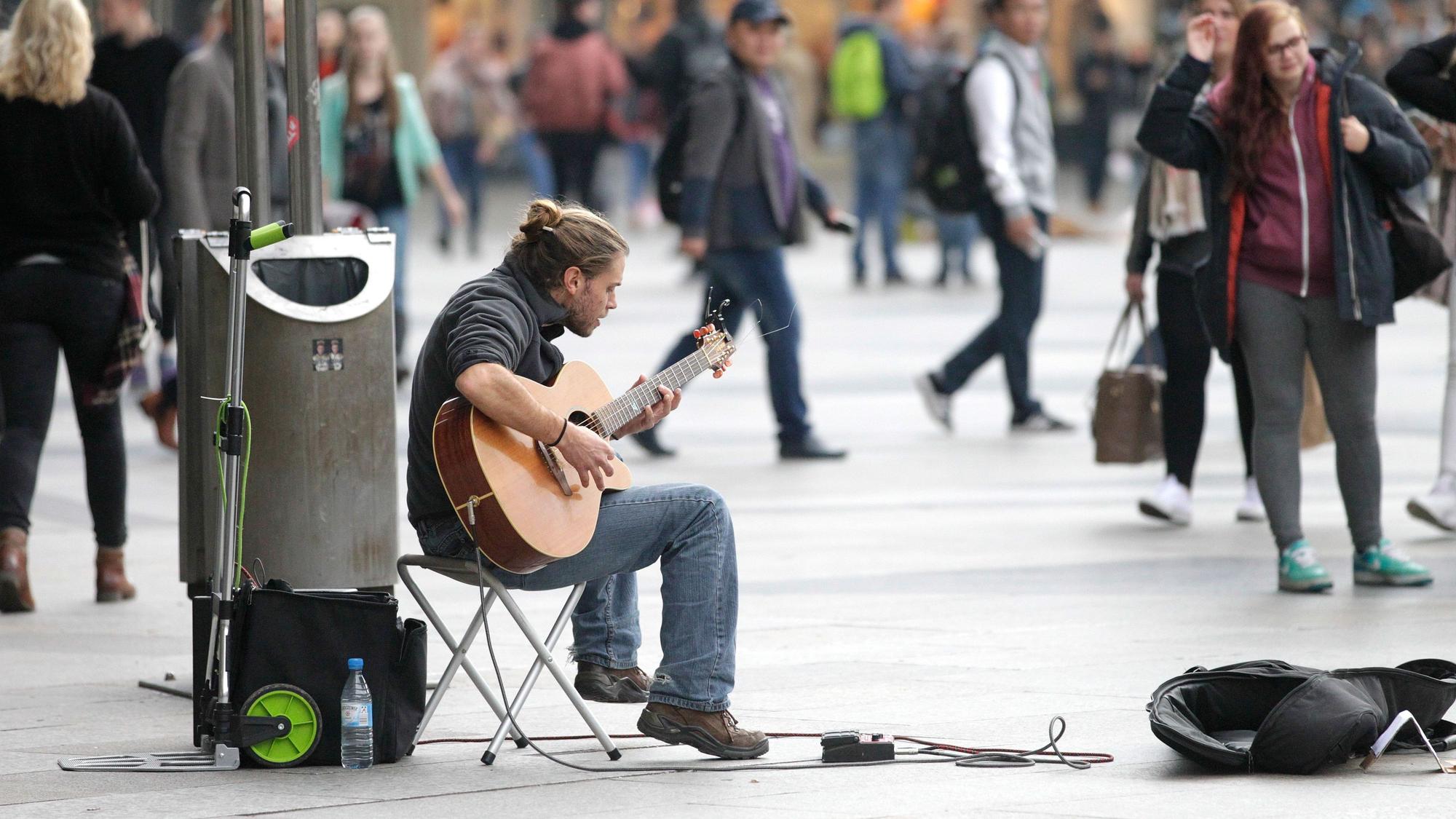 Ein junger Mann sitzt auf einem Klappstuhl auf der Kölner Domplatte und spielt Gitarre. Vor ihm liegt ein geöffneter Gitarrenkoffer, in den Passanten eine Anerkennung für sein Spiel legen können. Um ihn herum sind Passanten zu sehen, die ihrer Wege gehen. Eine Passantin hört ihm zu.