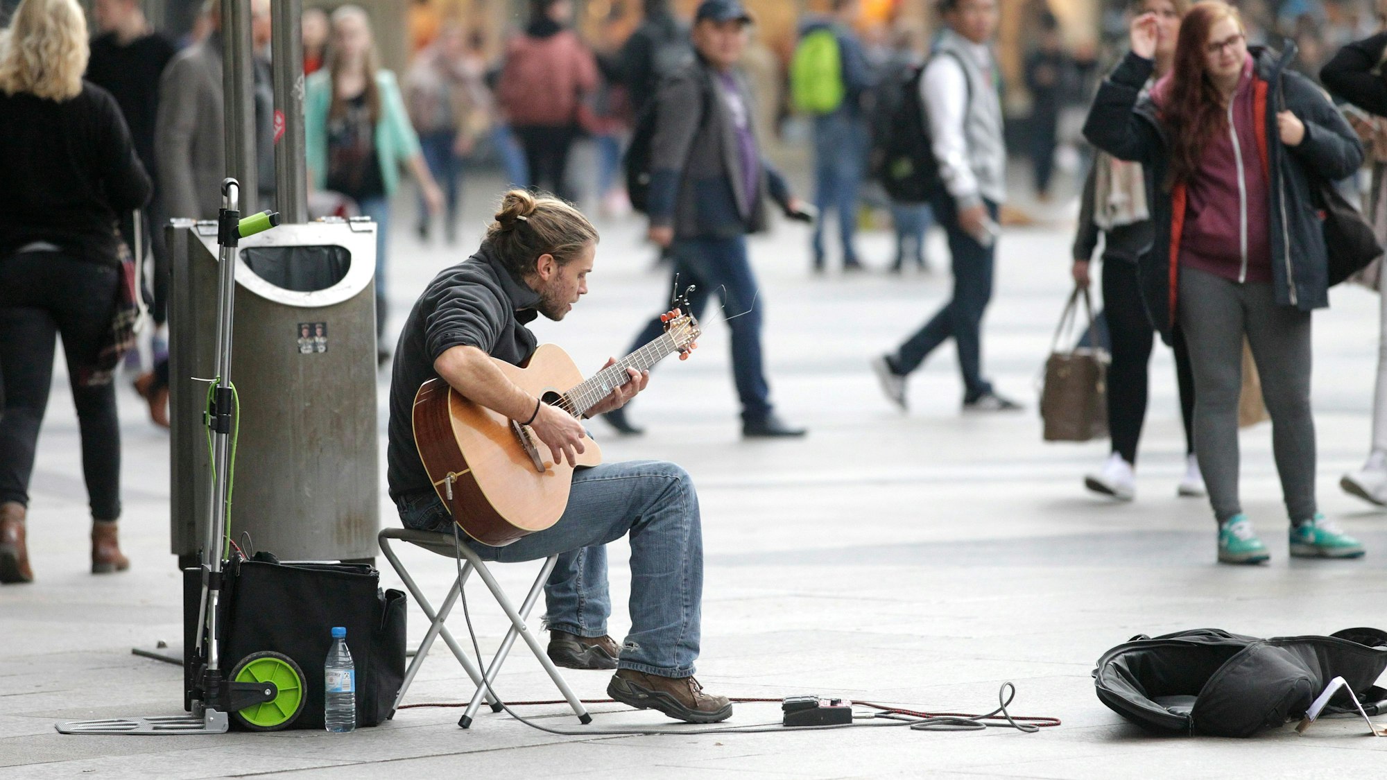 Ein junger Mann sitzt auf einem Klappstuhl auf der Kölner Domplatte und spielt Gitarre. Vor ihm liegt ein geöffneter Gitarrenkoffer, in den Passanten eine Anerkennung für sein Spiel legen können. Um ihn herum sind Passanten zu sehen, die ihrer Wege gehen. Eine Passantin hört ihm zu.