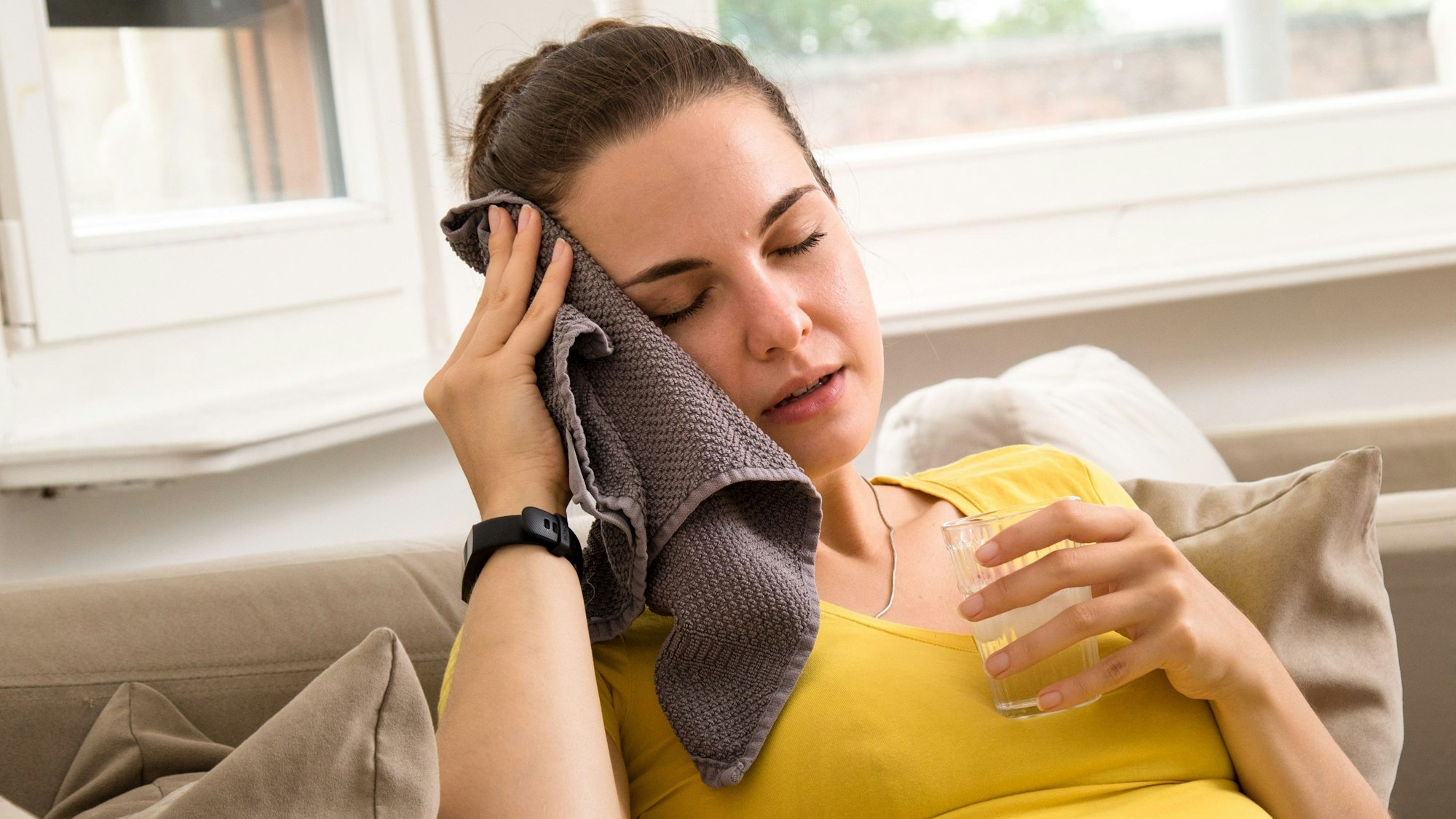 Eine Frau sitzt mit einem feuchten Tuch am Kopf und einem Glas Wasser in der Hand auf dem Sofa