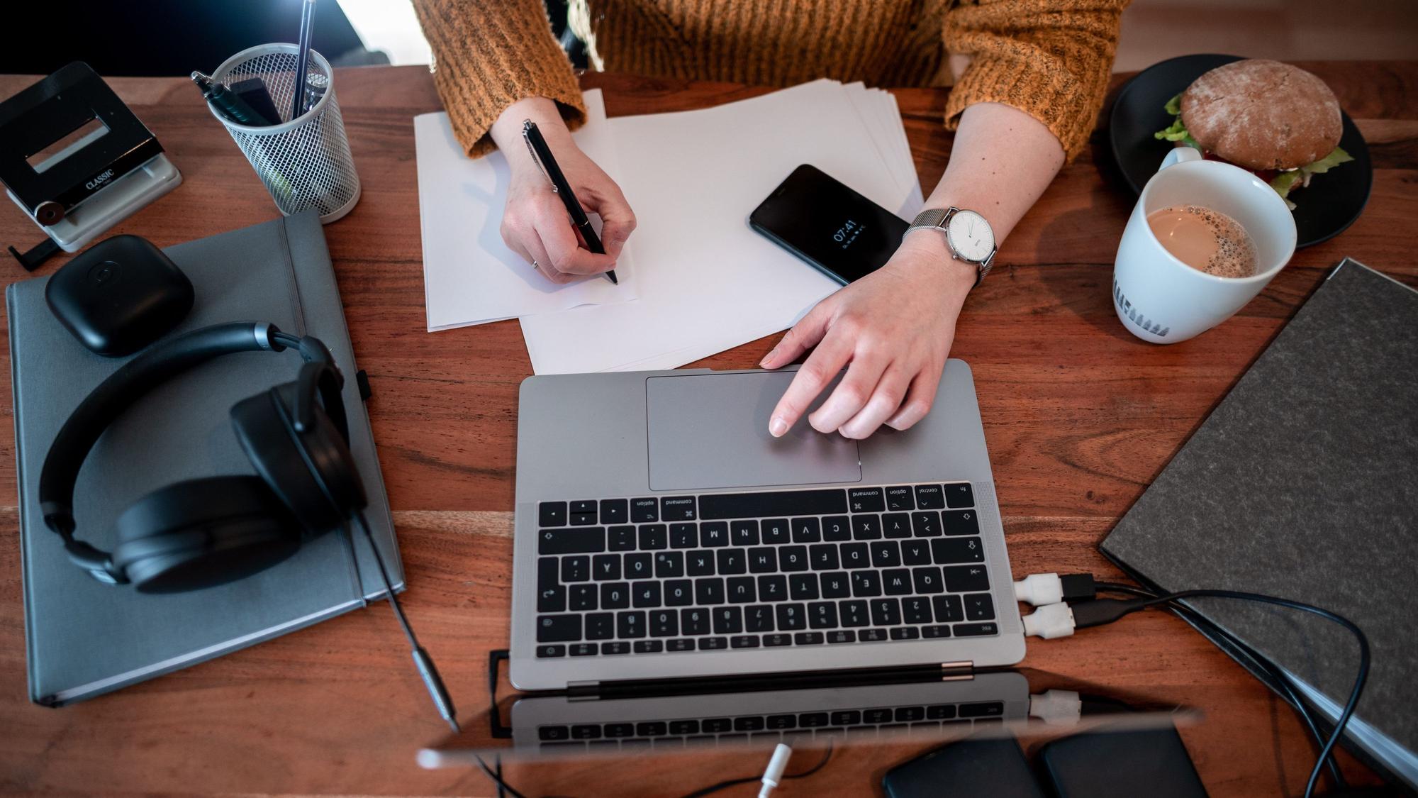 Eine Person sitzt an einem vollen Schreibtisch und arbeitet an einem Laptop