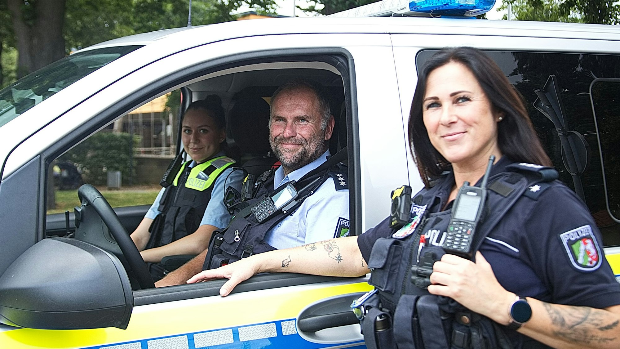 Das Bild zeigt zwei Polizeibeamtinnen und einen Polizeibeamten an einem Streifenwagen.