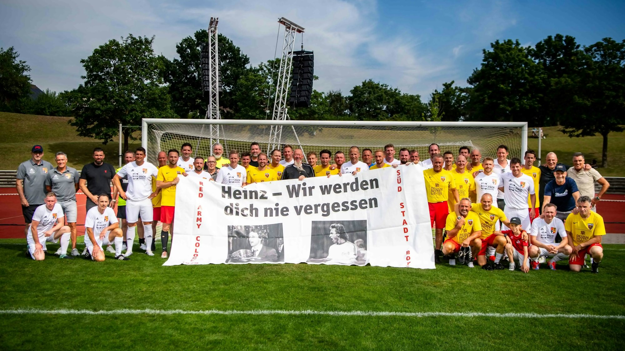 Die Spieler der beiden Teams haben sich vor einem Tor aufgestellt und halten ein großes Banner mit Fotos von Heinz Flohe und der Aufschrift „Heinz - Wir werden dich nie vergessen“.