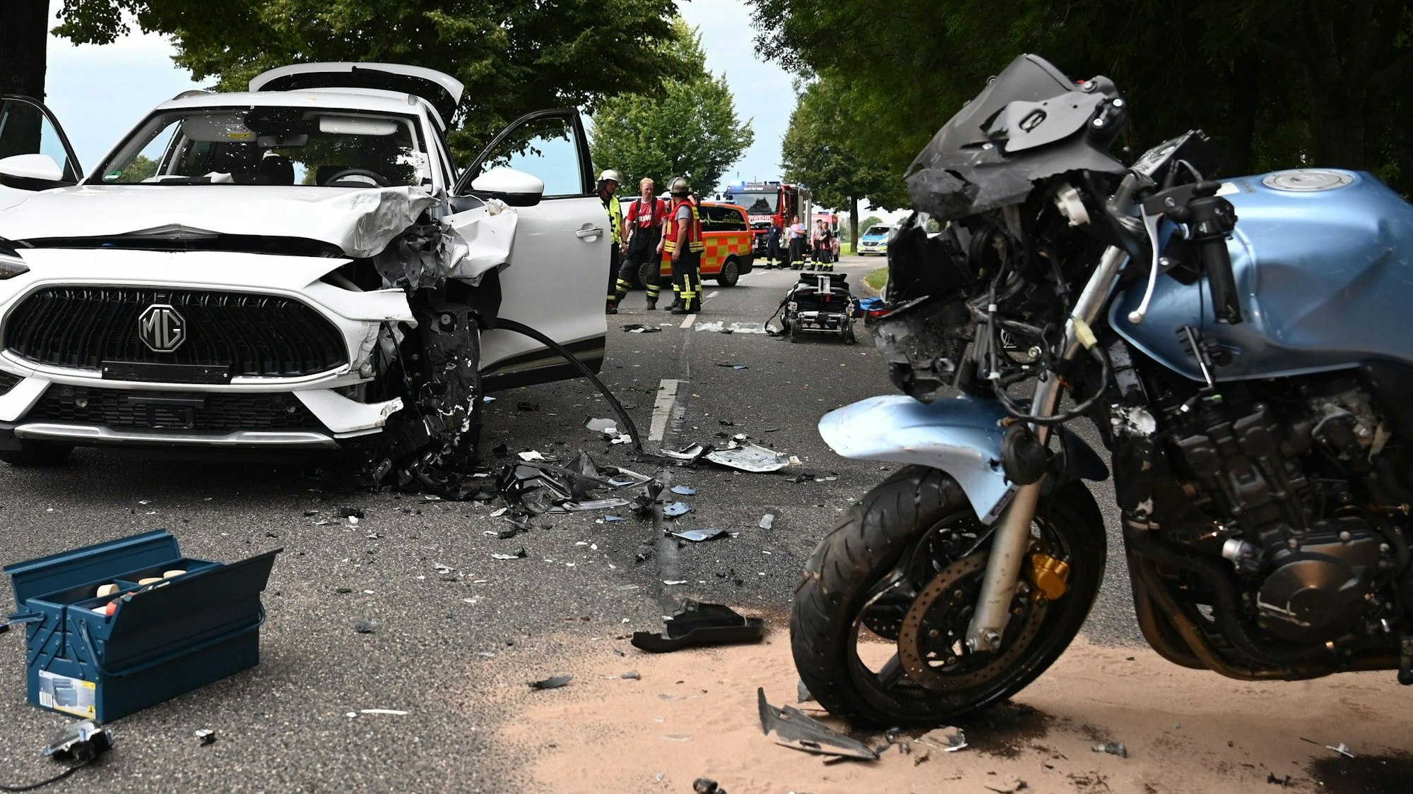 Das Bild zeigt die Unfallstelle in Elsdorf. Ein Motorrad und ein Auto stehen auf der Straße, mehrere Einsatzkräfte stehen an der Unfallstelle.