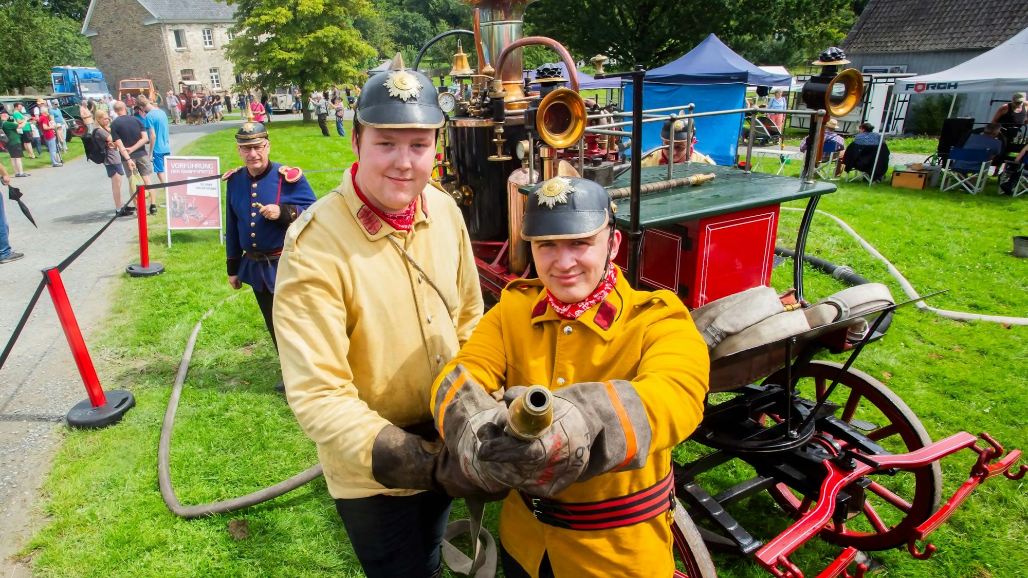 Zwei Männer in historischen Feuerwehr-Uniformen zielen mit einem Strahlrohr in die Kamera, hinter ihnen ist eine Dampfspritze aus dem Jahr 1901 zu erkennen.