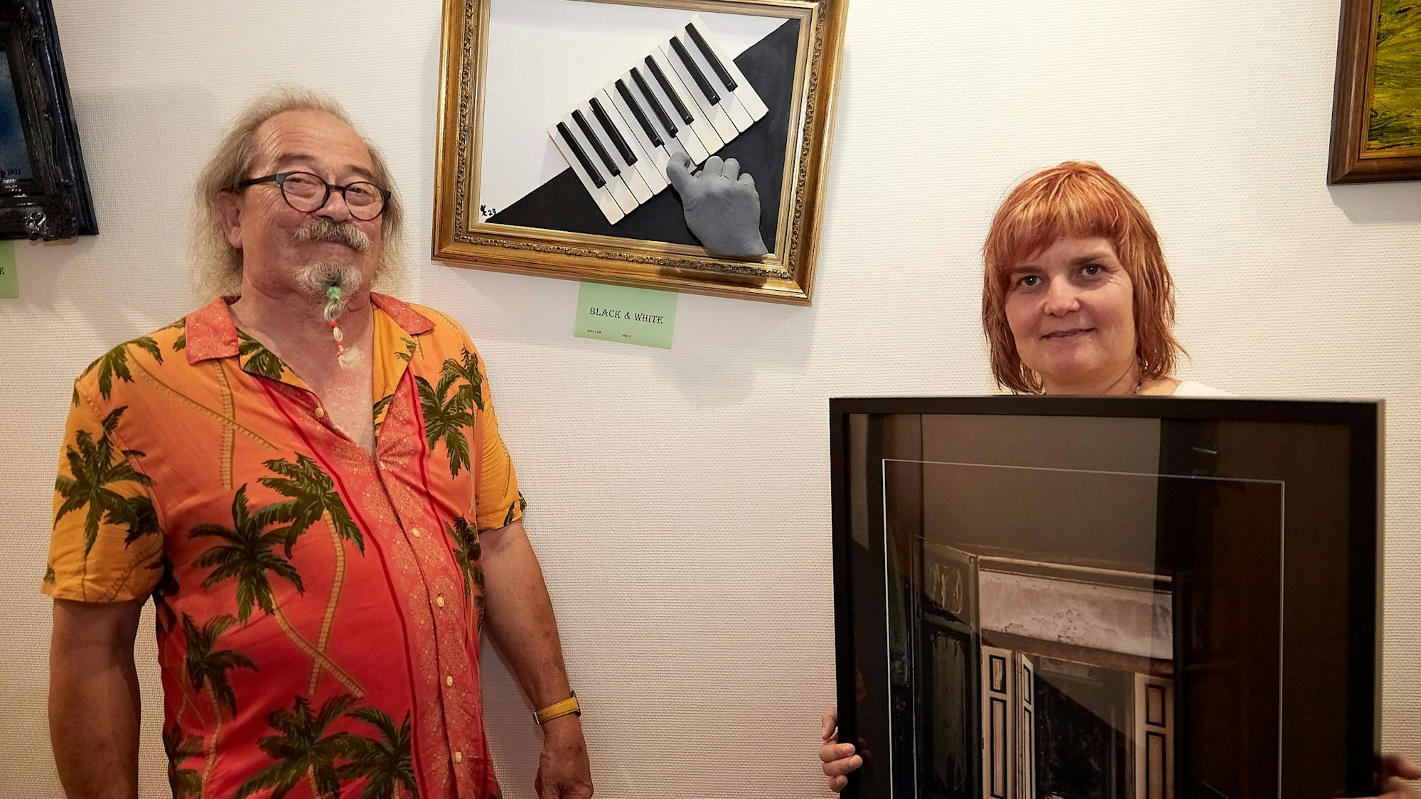 Das Foto zeigt Uwe Rhiem im bunten Hawaiihemd und Iris Hilgers, die eines ihrer Bilder in den Händen hält.