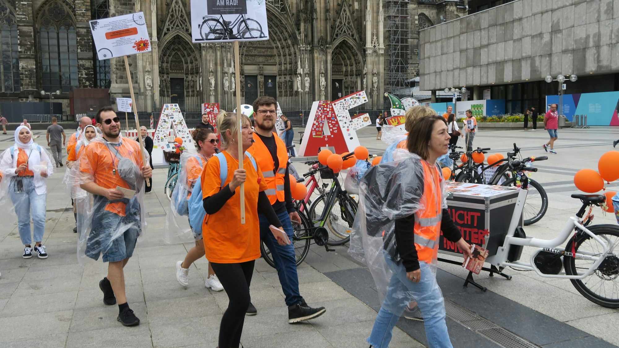 Männer und Frauen in orangen Oberteilen gehen über die Domplatte.
