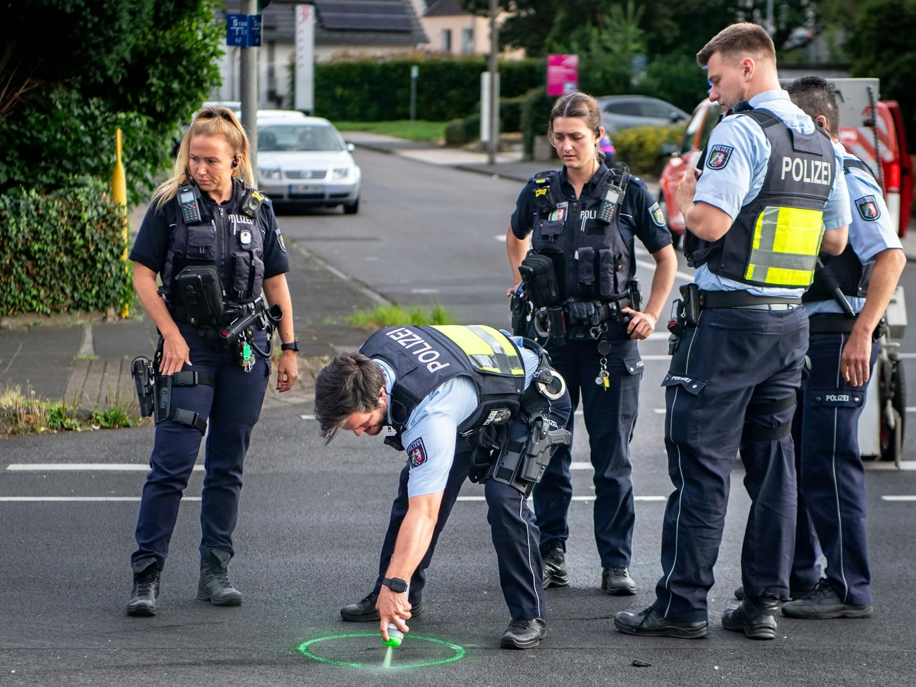 Mehrere Polizisten und Polizistinnen stehen in Uniform auf einer Straße. Ein Polizist sprüht einen grünen Kreis auf den Asphalt.