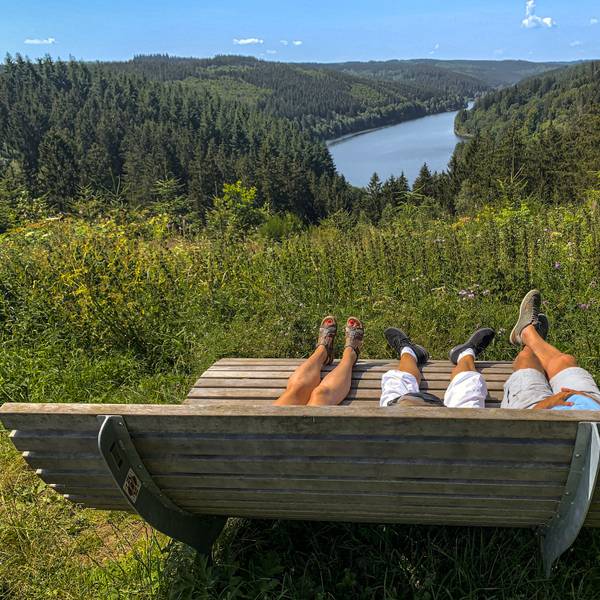 Auf einer von hinten fotografierten Ruhebank liegen drei Personen, von denen die Beine und Füße zu sehen sind. Dahinter befinden sich Wald und Talsperre.