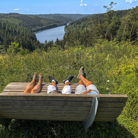 Auf einer von hinten fotografierten Ruhebank liegen drei Personen, von denen die Beine und Füße zu sehen sind. Dahinter befinden sich Wald und Talsperre.
