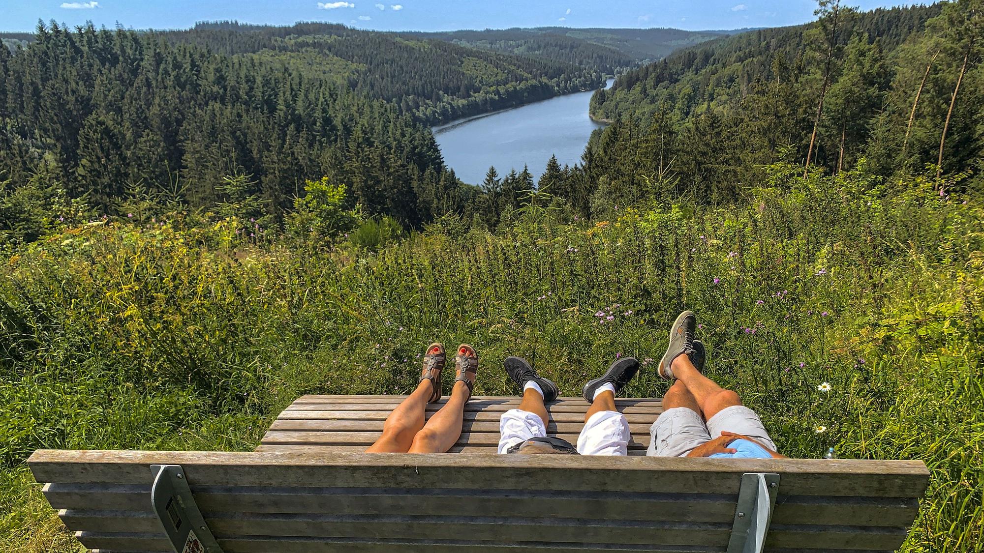 Auf einer von hinten fotografierten Ruhebank liegen drei Personen, von denen die Beine und Füße zu sehen sind. Dahinter befinden sich Wald und Talsperre.