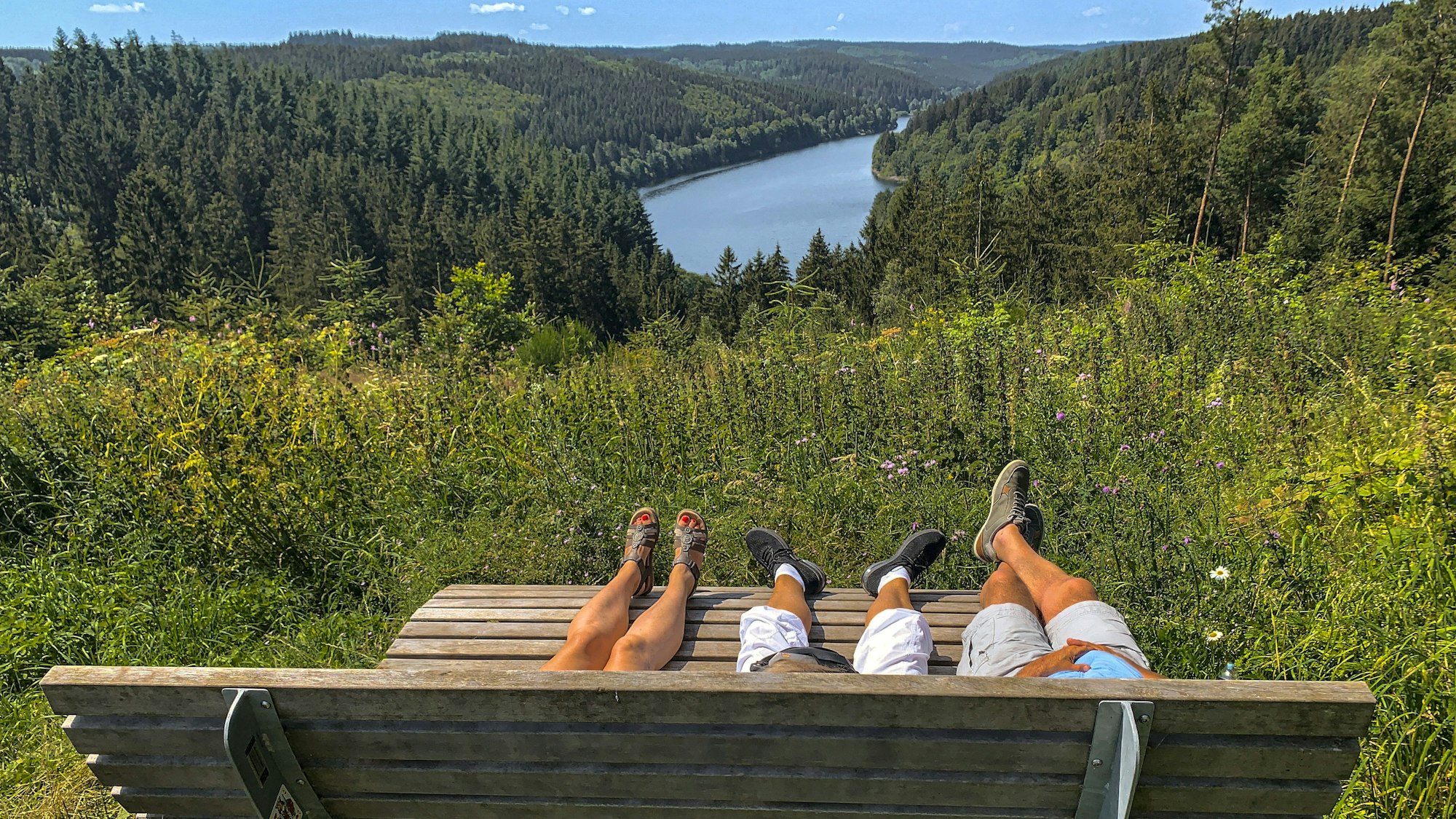 Auf einer von hinten fotografierten Ruhebank liegen drei Personen, von denen die Beine und Füße zu sehen sind. Dahinter befinden sich Wald und Talsperre.