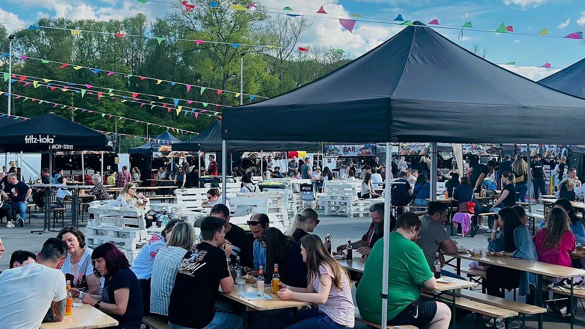 Menschen sitzen unter Pavillons an Bierzeltgarnituren und essen.