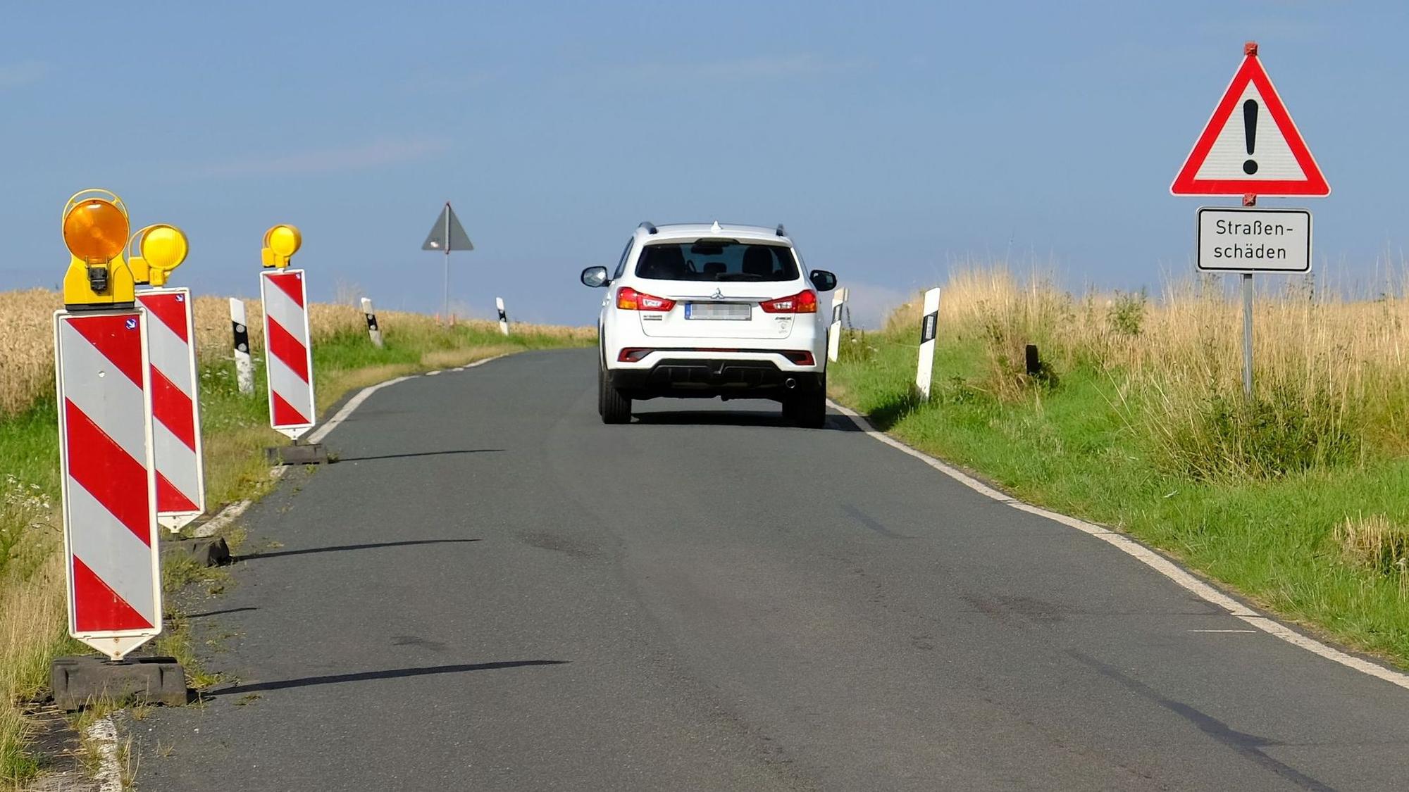 Ein Warnschild weist auf der K58 auf bestehende Straßenschäden hin; ein weißes SUV ist von hinten zu sehen.