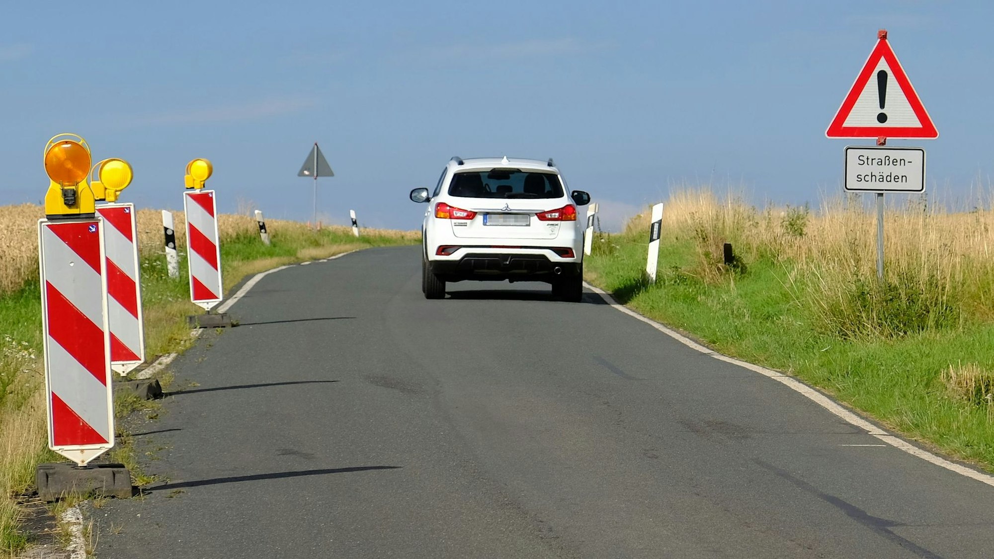 Ein Warnschild weist auf der K58 auf bestehende Straßenschäden hin; ein weißes SUV ist von hinten zu sehen.