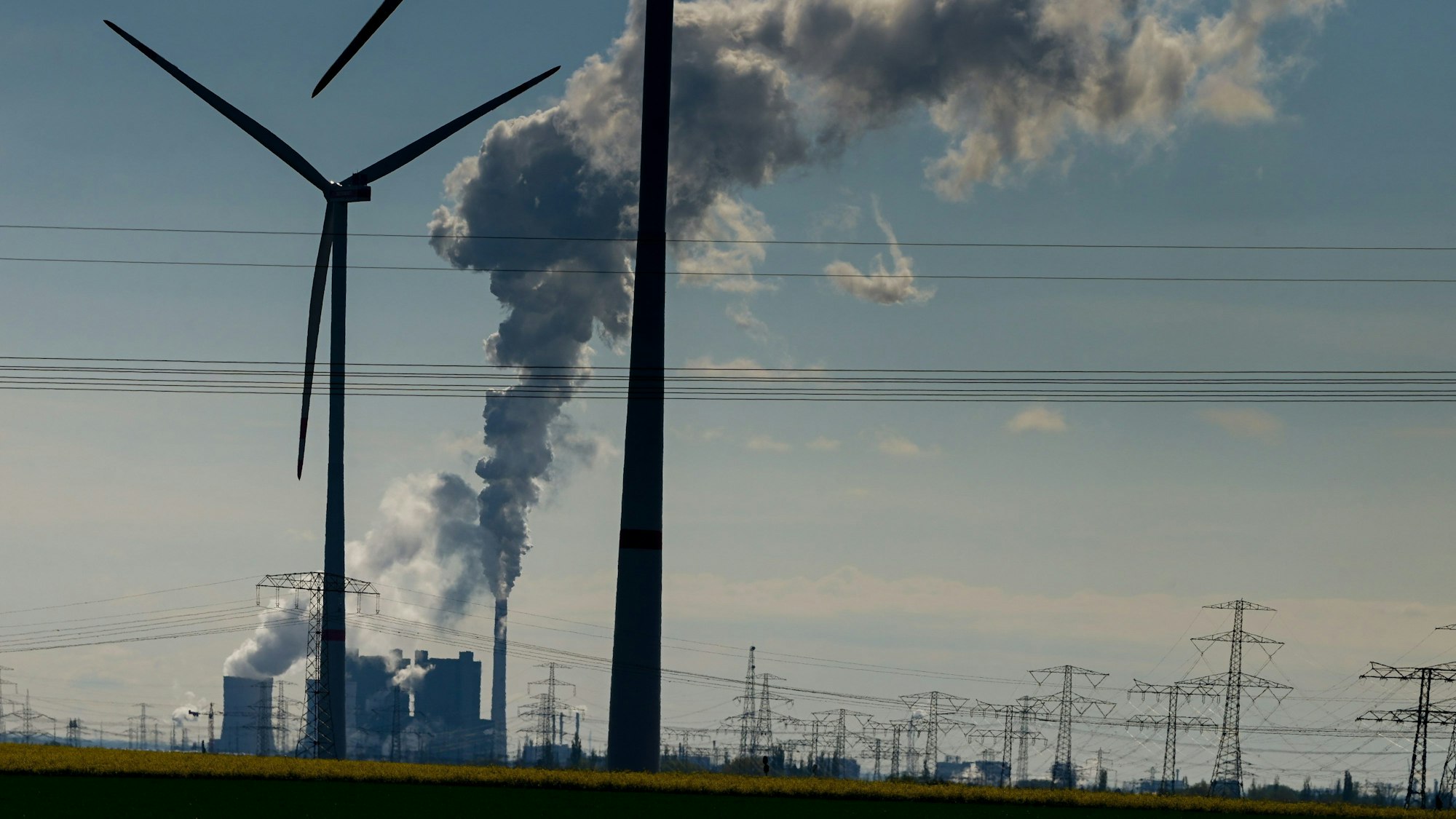 Windräder stehen auf einem Feld.Im Hintergrund rauchen Schornsteine eines Industriegebiets, umgeben von einer Stromtrasse.