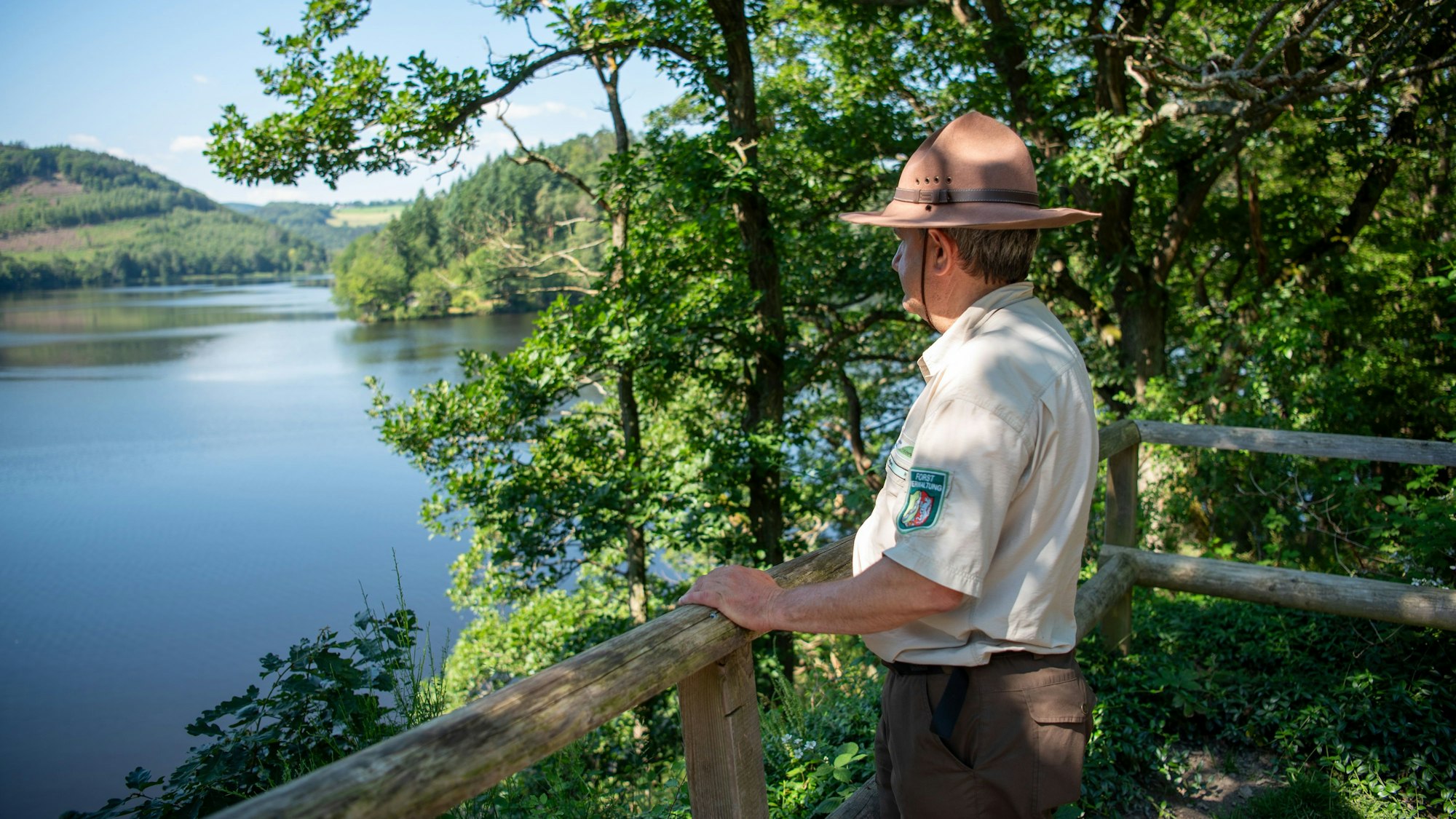 Ranger Ralf Hilgers steht in dunkelbrauner Hose, beigem Hemd und braunem Rangerhut an einem Holzgeländer und schaut auf den Rursee.