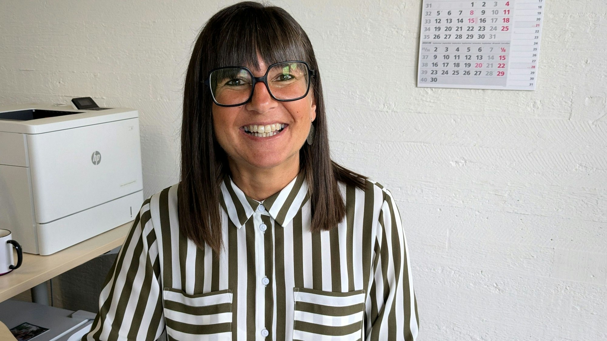 Susanne Aleksander in ihrem Büro: links steht ein Drucker, an der Wand hängt ein Dreimonatskalender. Sie trägt eine gestreifte Bluse und eine Brille.