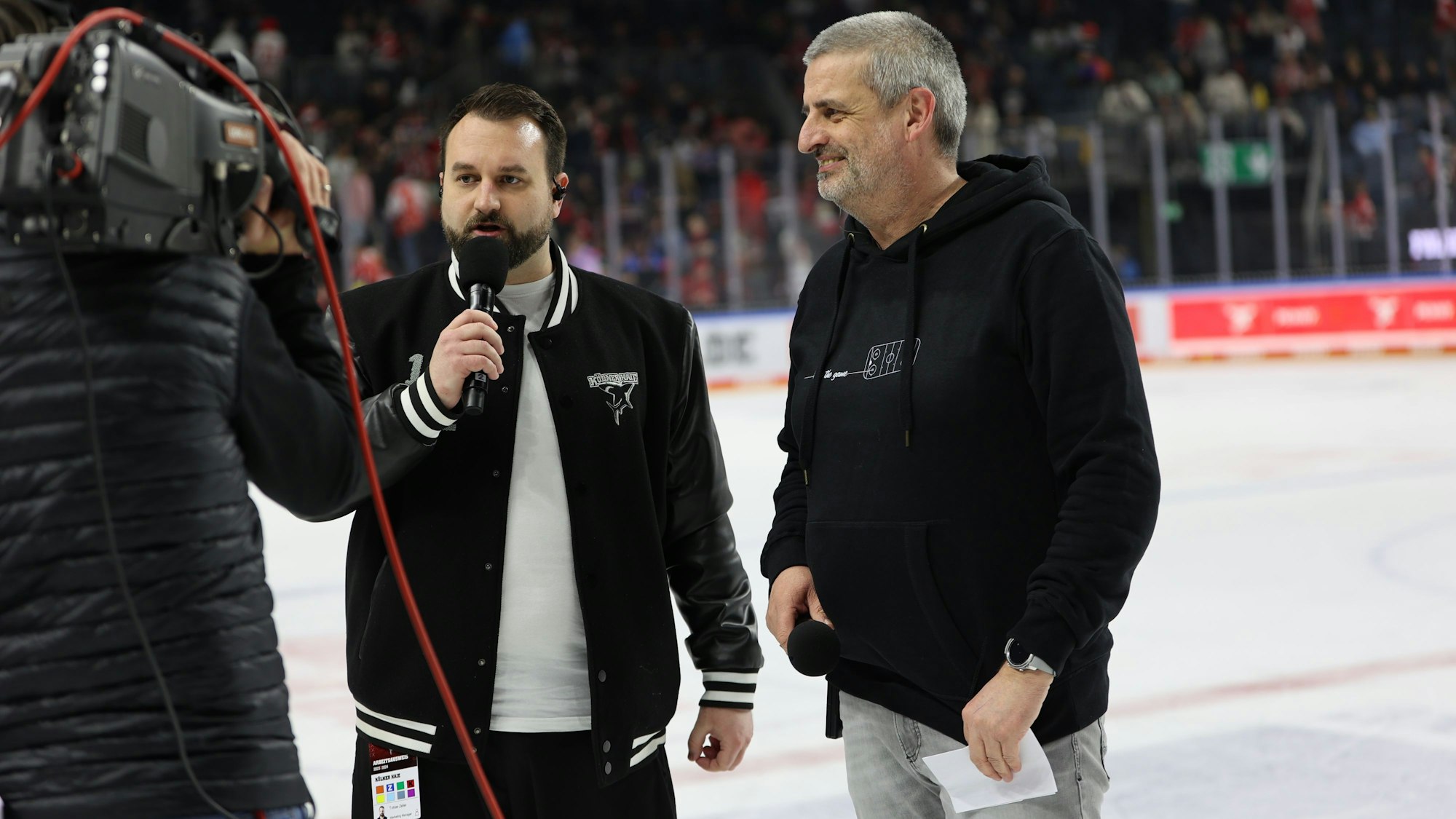 Zwei Männer stehen auf der Eisfläche eines Eishockeyfeldes. Einer hält ein Mikrofon in der Hand. Links ist ein Kameramann von hinten zu sehen.