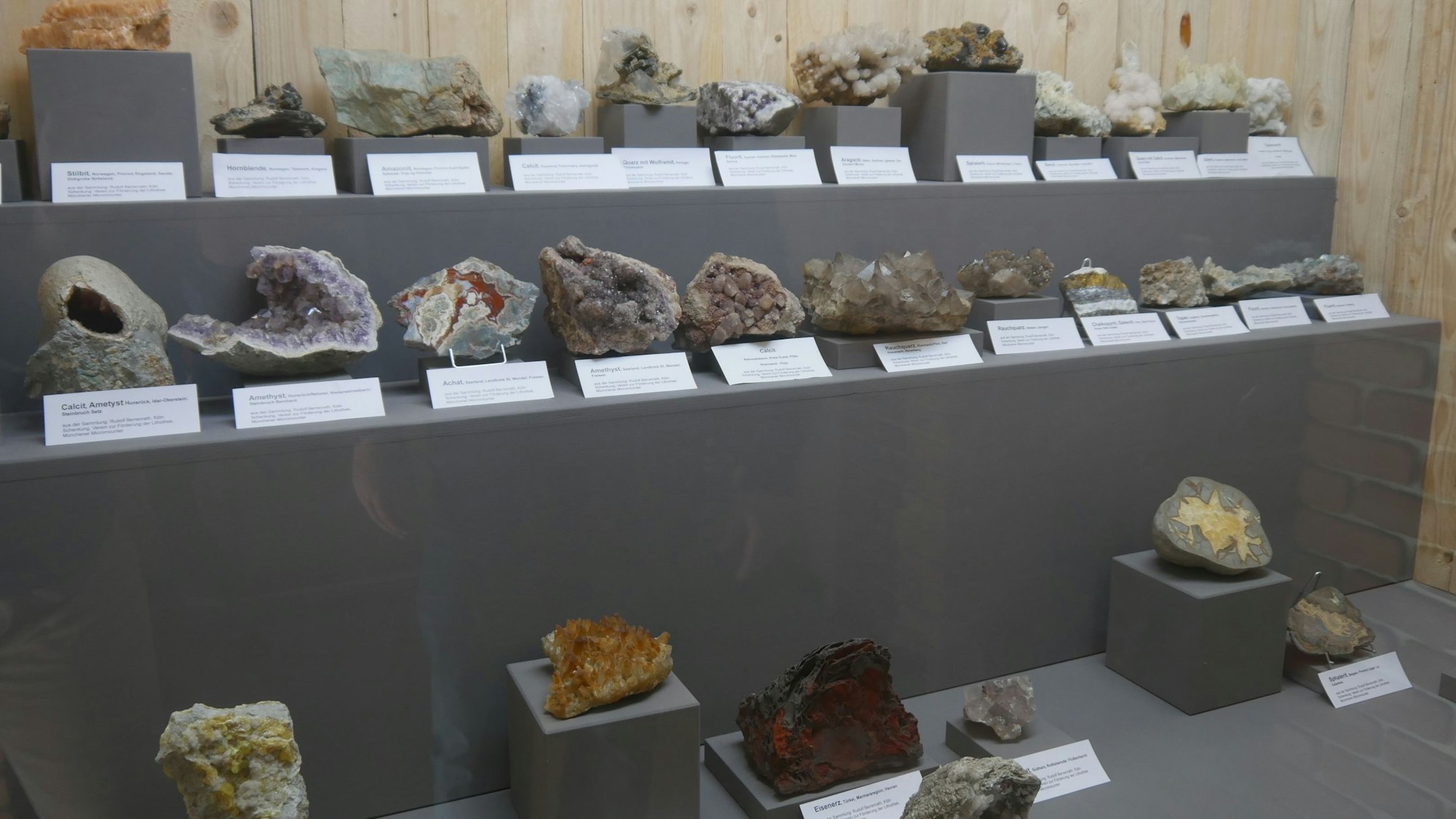 Das Bild zeigt 30 verschiedenfarbige Mineralien mit bizarren Formen in einer Ausstellung.