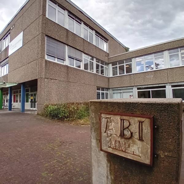 Blick auf das Schulgebäude, im Vordergrund ein Betonblock mit verwitterter Inschrift "Abi".
