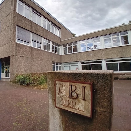 Blick auf das Schulgebäude, im Vordergrund ein Betonblock mit verwitterter Inschrift "Abi".