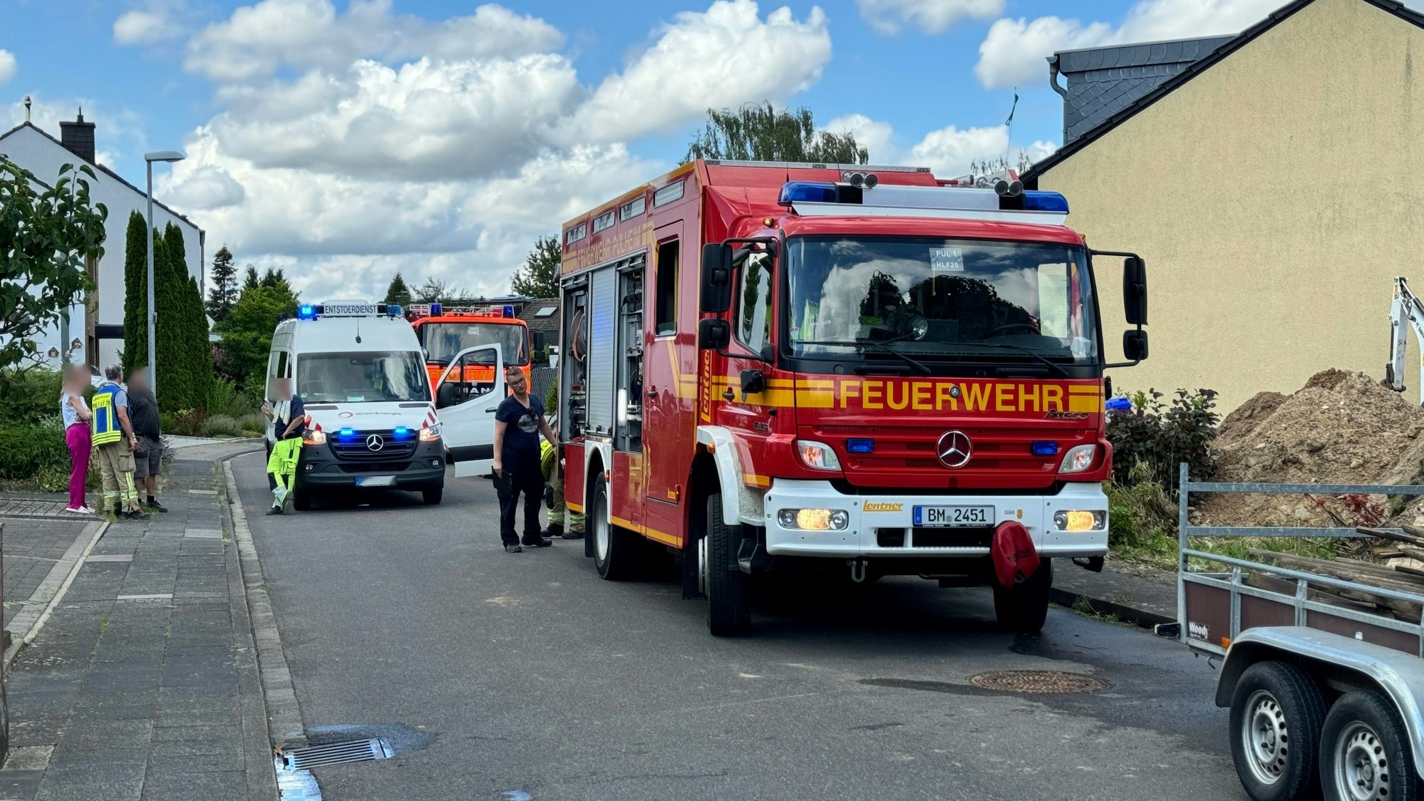 Ein Feuerwehrfahrzeug, mehrere Personen und Einsatzkräfte. Rechts im Bild ein Sandhaufen und der Arm eines kleinen Baggers.
