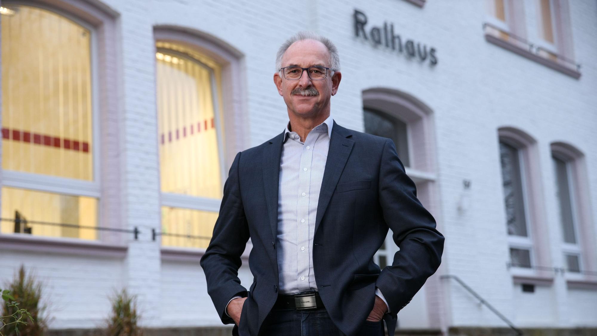 Ein Mann im Anzug, mit Brille und Schnurrbart steht vor einem weißen Gebäude. Über dem Eingang steht "Rathaus".
