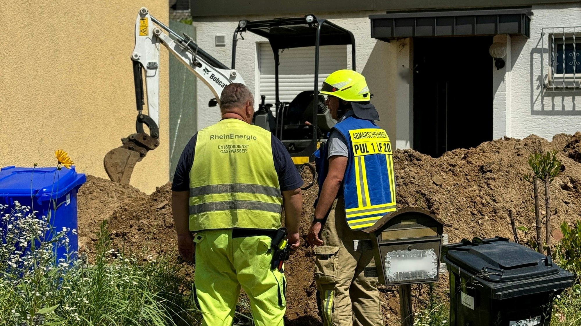 Zwei Einsatzkräfte der Feuerwehr. Im Hintergrund der Arm eines Mini-Baggers an einer Hauswand.