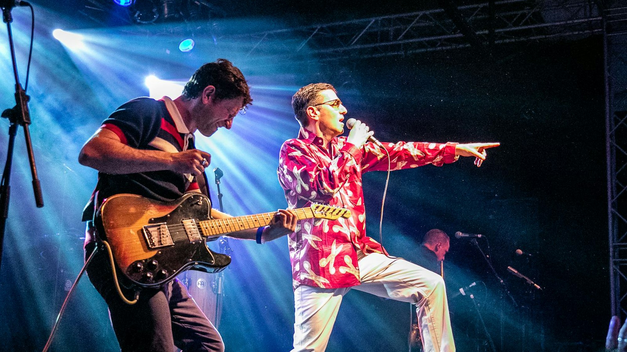 Zu sehen sind ein Gitarrist und ein Sänger, die auf einer Bühne performen.