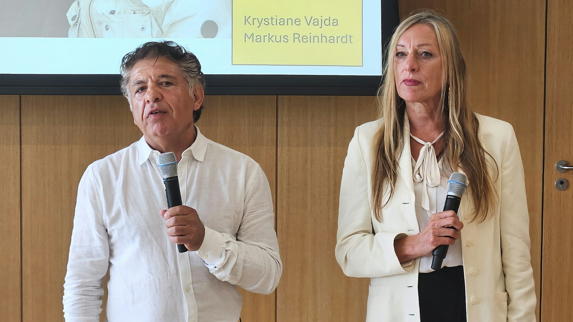 Markus Reinhardt und Krystiane Vajda stehen mit Mikros in der Hand nebeneinander.