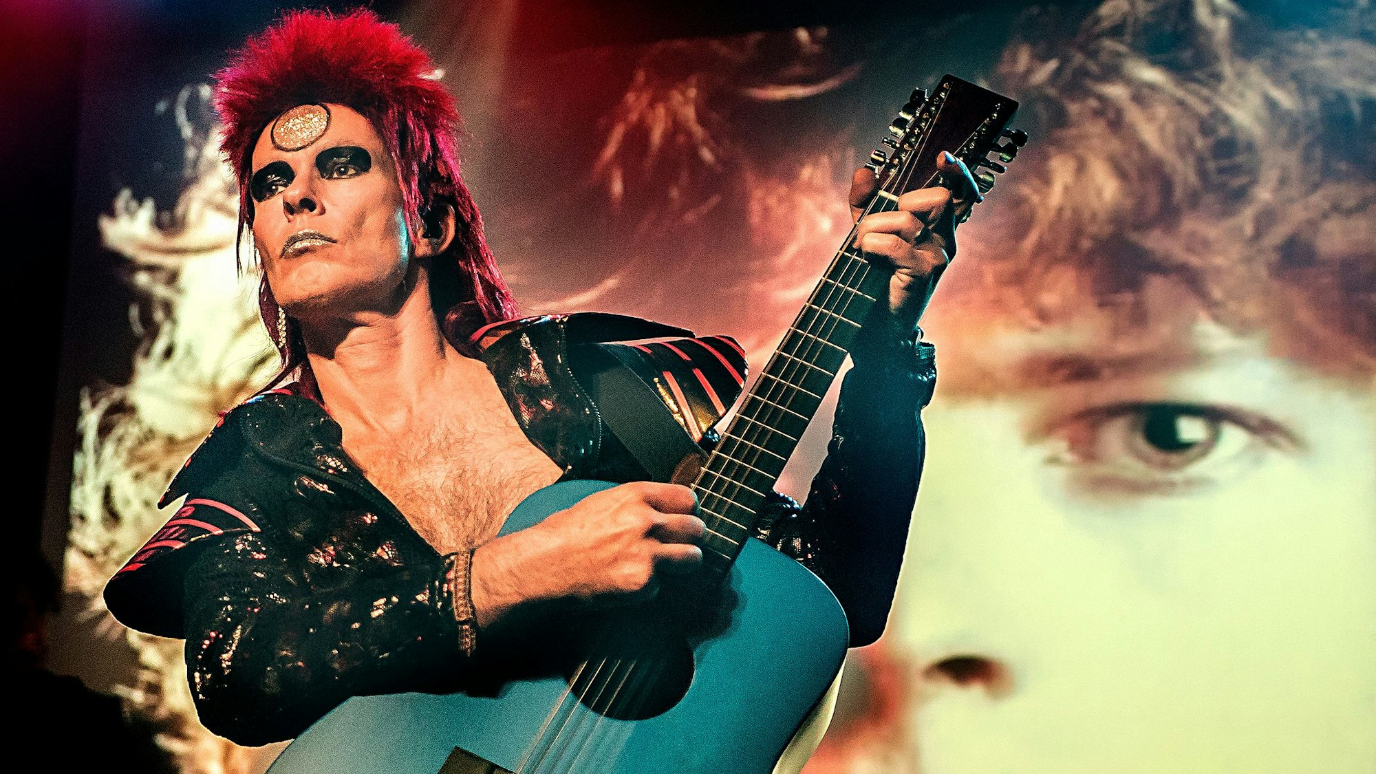 Zu sehen ist ein Musiker, der auf einer Bühne mit einer Gitarre performt.