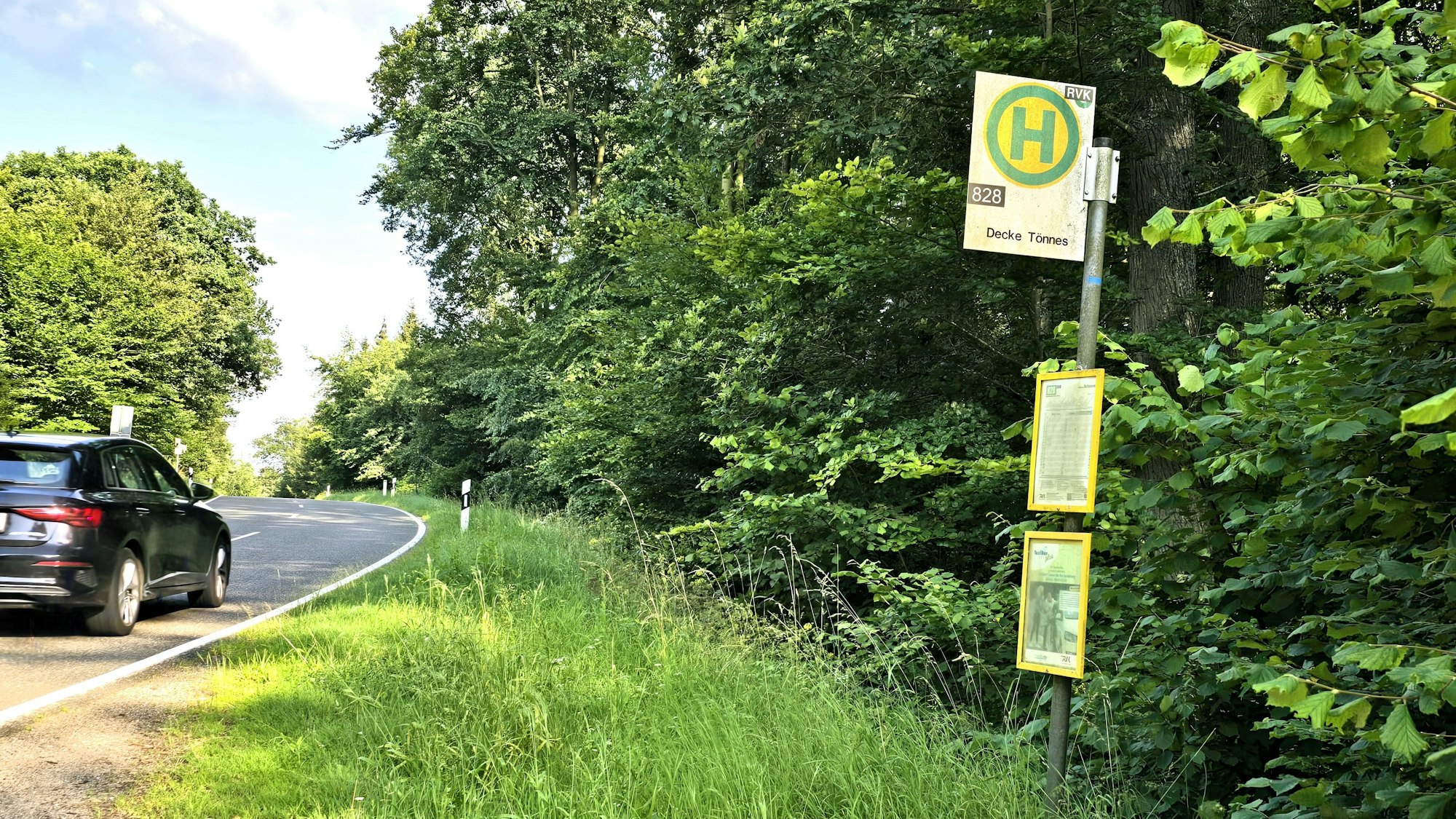 Nur wenige Meter von der bekannten Waldkapelle Decke Tönnes zwischen Effelsberg und Bad Münstereifel entfernt, steht ein Bushalteschild an einer Landstraße. Rechts im Bild die Haltestelle, mittig Wiese, links im Bild fährt ein Auto über die asphaltierte Straße.
