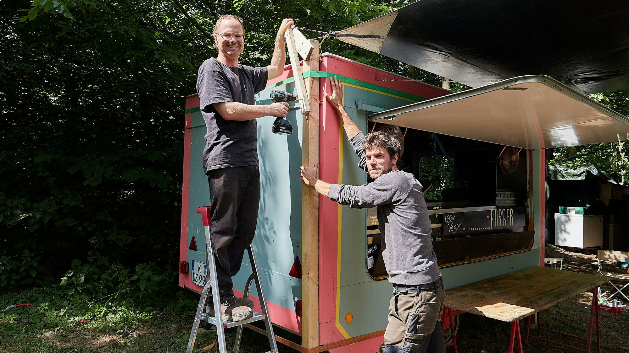 Zwei Männer, Timon Verleger (links) und Björn Altenburg, bauen eine Imbissbude auf, deren Lade schon hochgeklappt ist. Hier werden sie beim Zugvögel-Festival am Weißen Stein in Hellenthal-Udenbreth vegane Burger anbieten.