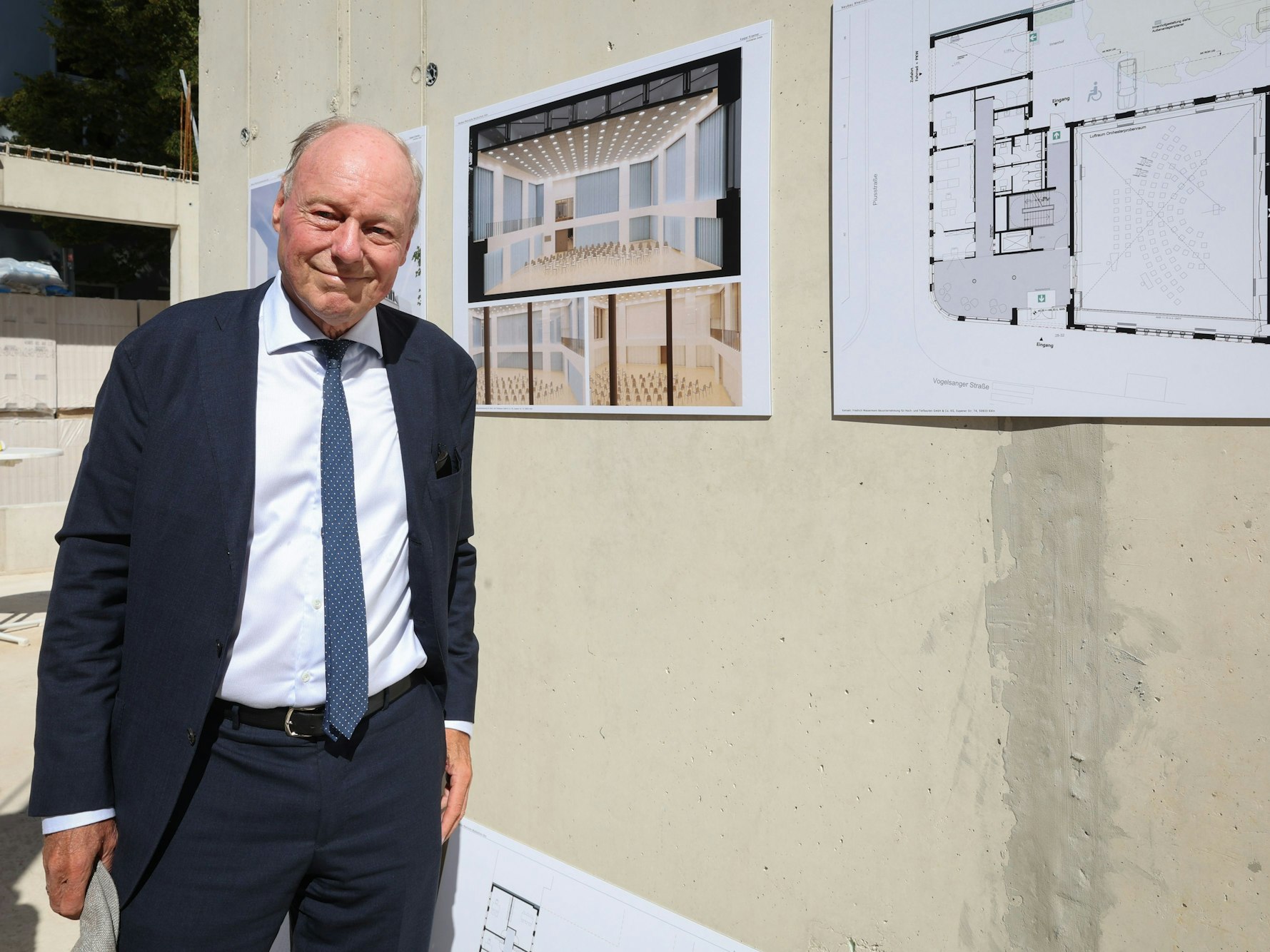 Der Architekt Kaspar Kraemer
posiert lächelnd vor Entwurfsskizzen und Visualisierungen an einer Wand.