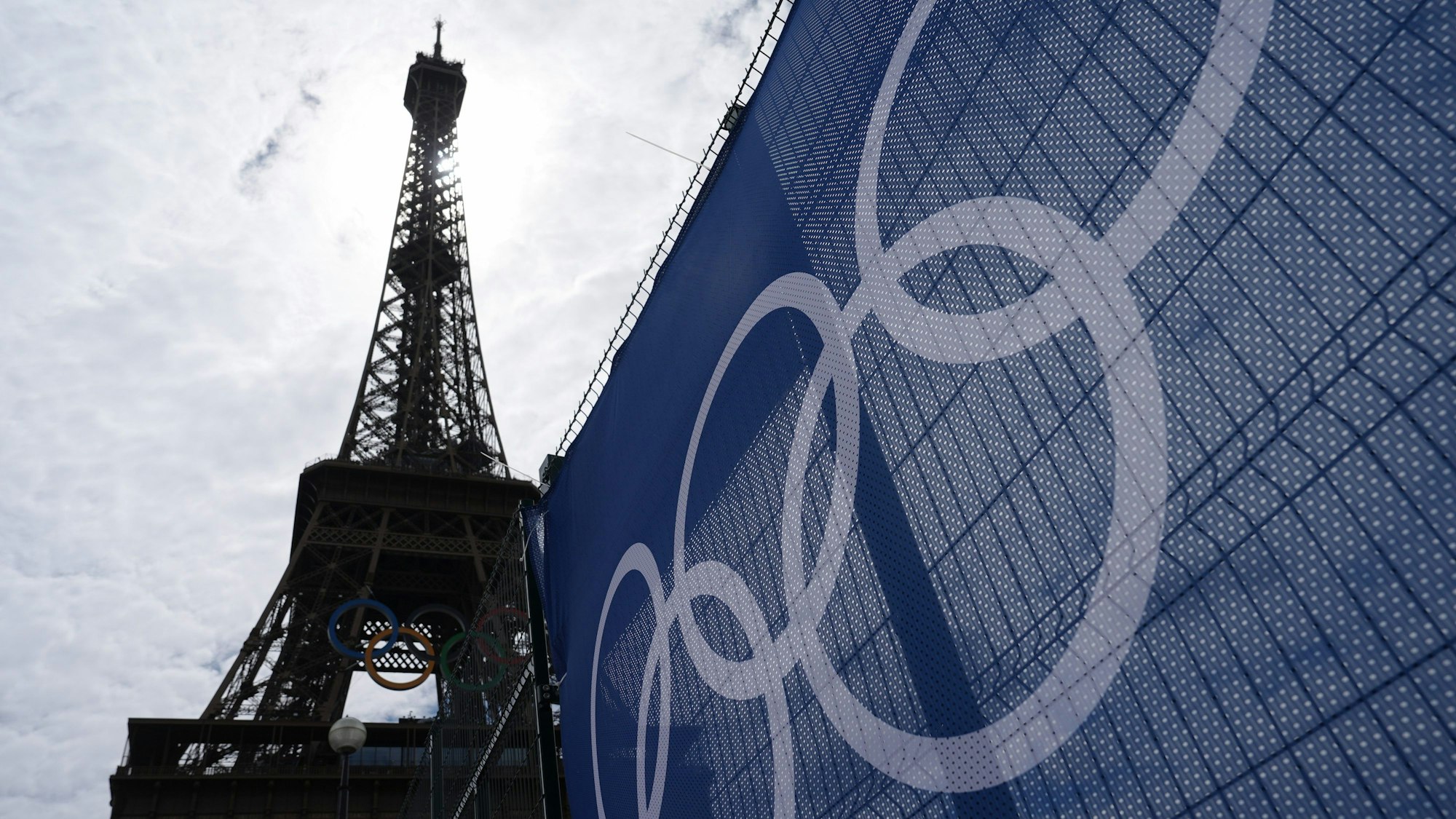 25.07.2024, Frankreich, Paris: Olympia, Olympischen Sommerspiele, Paris 2024, Vorbereitungen, der Eiffelturm im Gegenlicht neben einer Plane mit den Olympischen Ringen.