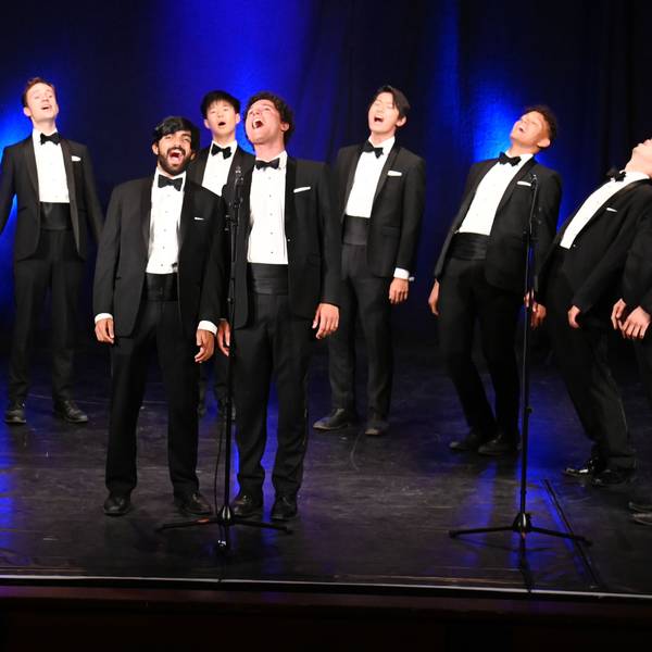 Zu sehen sind neun Männer in Anzügen auf einer Bühne, die singen.