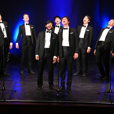 Zu sehen sind neun Männer in Anzügen auf einer Bühne, die singen.