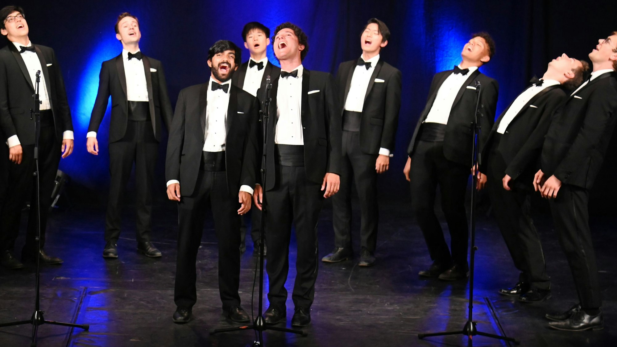 Zu sehen sind neun Männer in Anzügen auf einer Bühne, die singen.