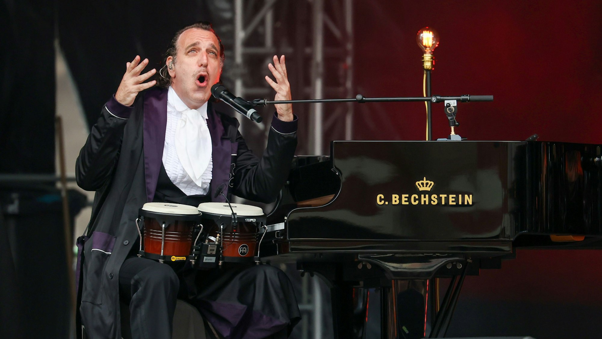 Chilly Gonzales sitzt mit Rüschenhemd und schwarzem Morgenmantel am Flügel, auf dem Schoß zwei Bongo-Trommeln. Er gestikuliert mit den Händen, während er singt. 25.07.2024
Köln:
Roncalliplatz Konzerte:
Pianist Chilly Gonzales
Foto: Martina Goyert