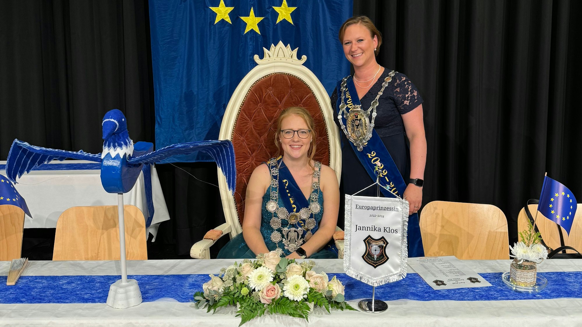 Zwei Frauen, eine sitzt, die andere steht an ihrer Seite, hinter ihnen hängt die Europafahne.