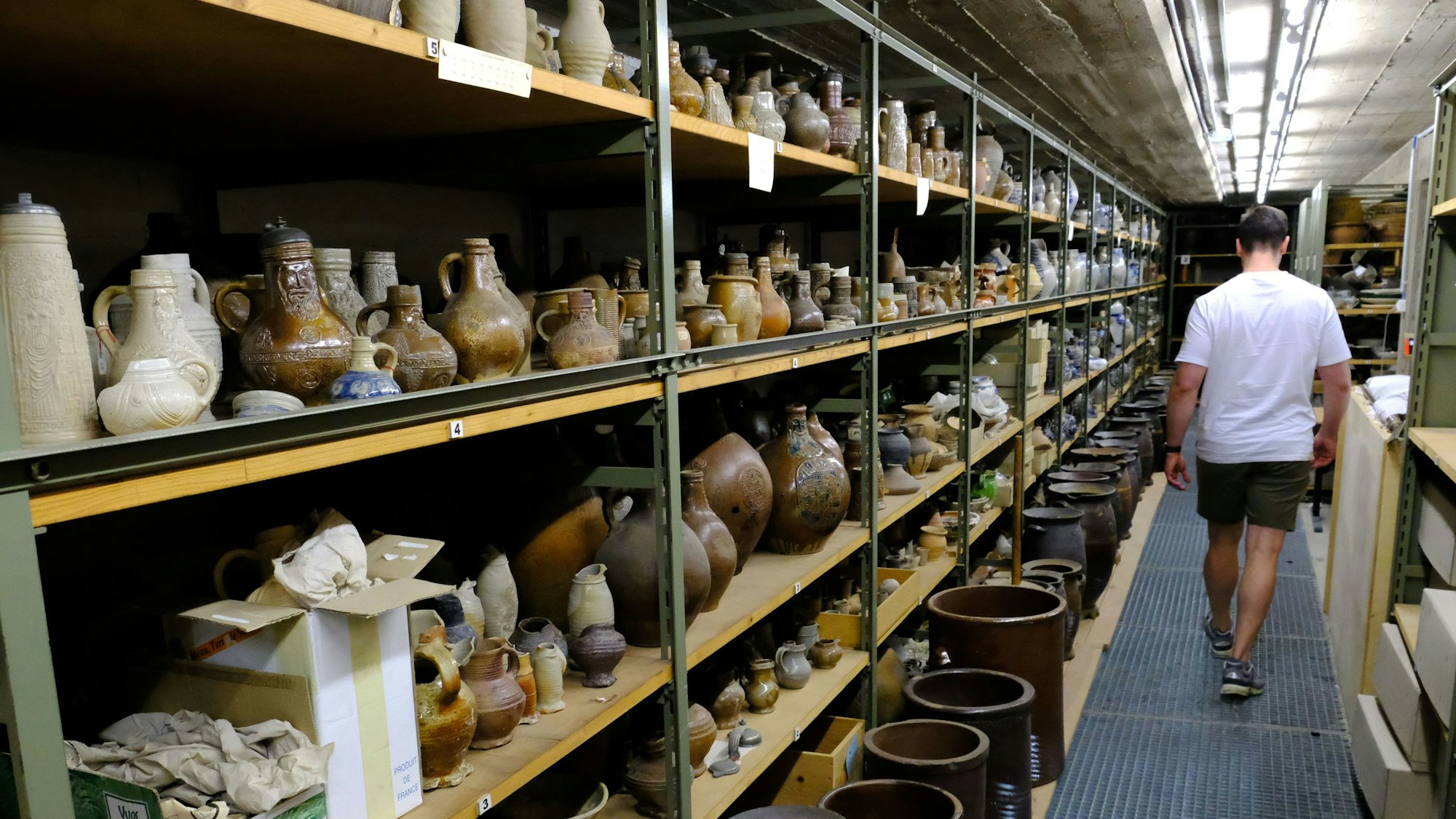 Töpferwaren aus dem Rheinland lagern in Regalen des Kommerner Museums-Depots. Ein Mitarbeiter geht daran vorbei.