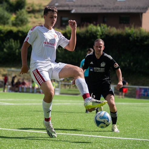 Ein junger Mann in einem weißen Fußballtrikot schaut auf den Fußball, der über den Rasen des Fußballplatzes in seine Richtung fliegt.