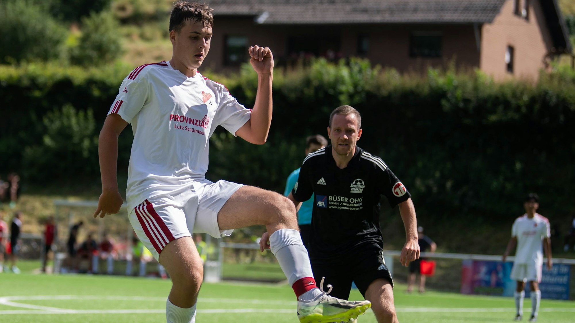 Ein junger Mann in einem weißen Fußballtrikot schaut auf den Fußball, der über den Rasen des Fußballplatzes in seine Richtung fliegt.