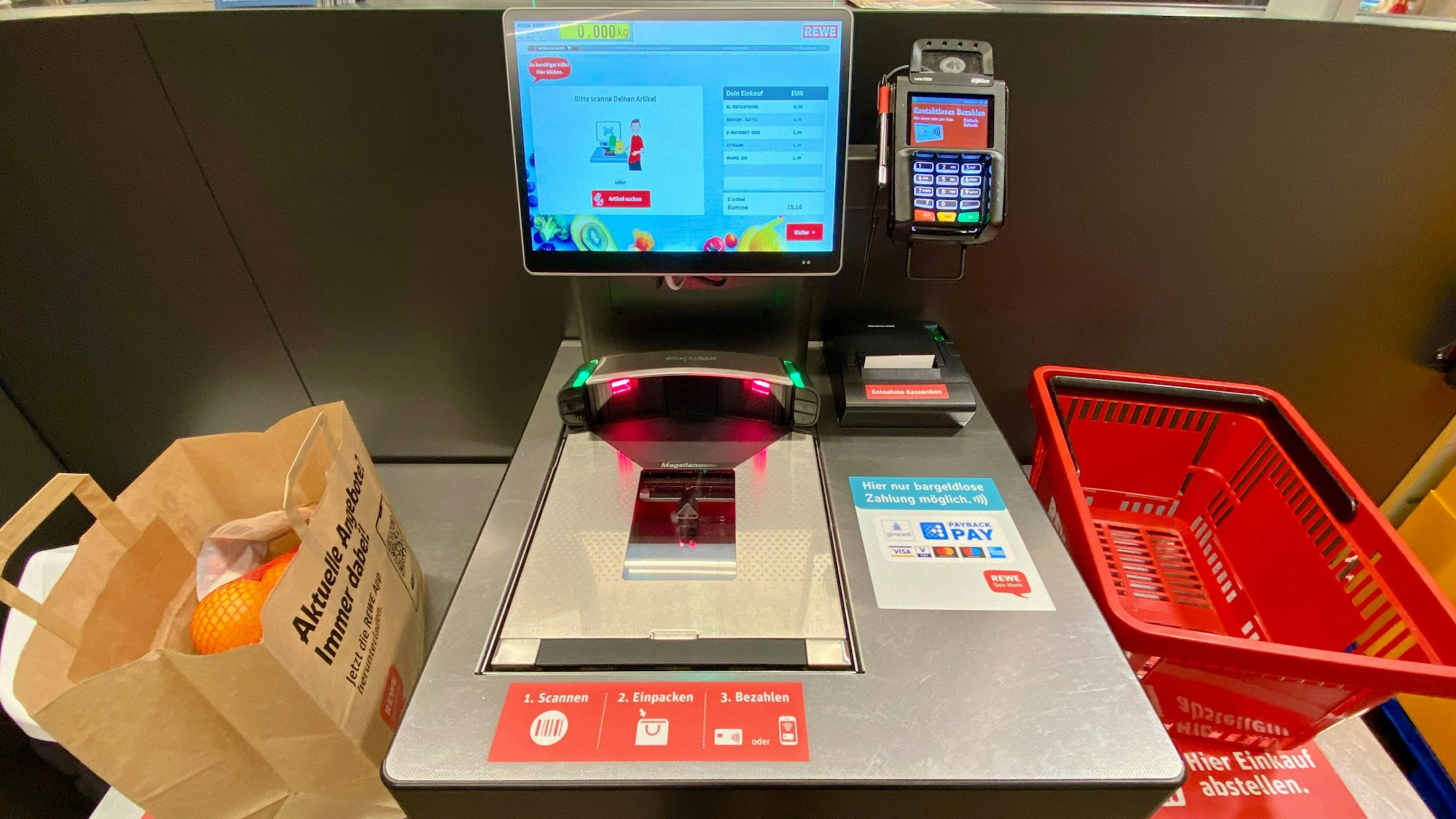 Das Foto zeig eine Selbstbedienungskasse, die ohne Kassierer auskommt. Sie besteht aus einem Scanner, einem Touchdisplay zur Preisermittlung von unverpacktem Obst und Gemüse sowie einem EC-Karten-Lesegerät zum bargeldlosen Bezahlen. Rechts der Kasse steht ein leerer Einkaufskorb, links eine mit Lebensmitteln gefüllte Papiertüte.
