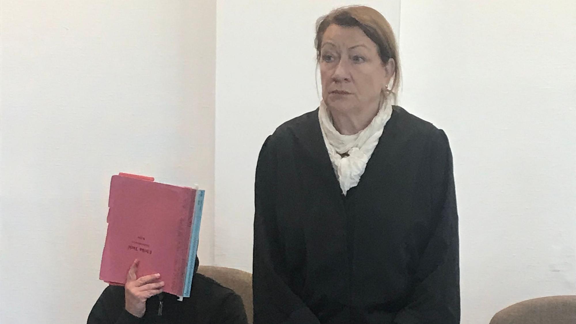 Zu sehen sind ein Angeklagter, der sein Gesicht mit einer roten Mappe verdeckt, und seine Verteidigerin in einem Gerichtssaal.