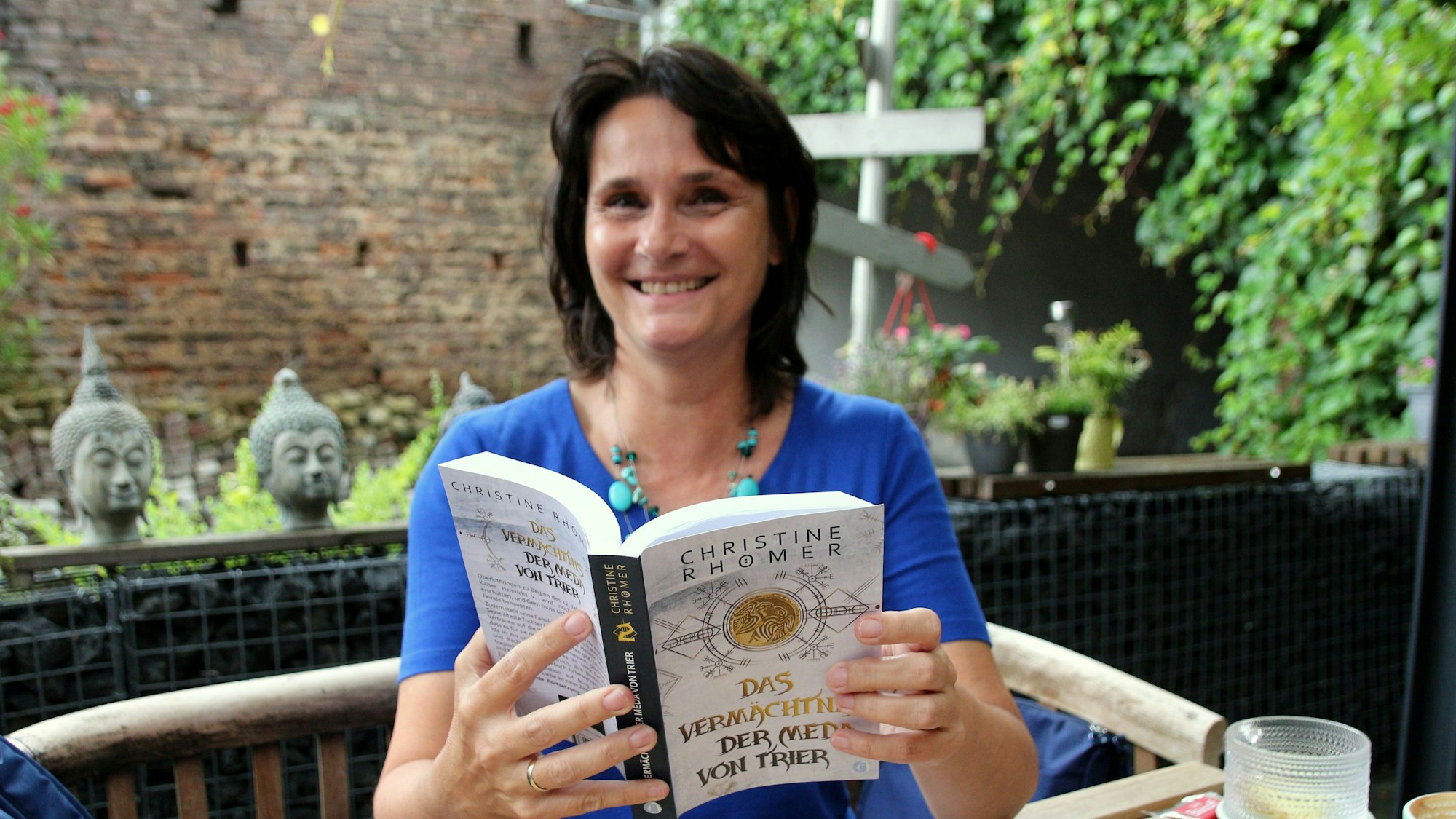 Eine Frau sitzt an einem Tisch in ihrem Garten, in ihren Händen hält sie ihr Buch, das in wenigen Tagen erscheint.