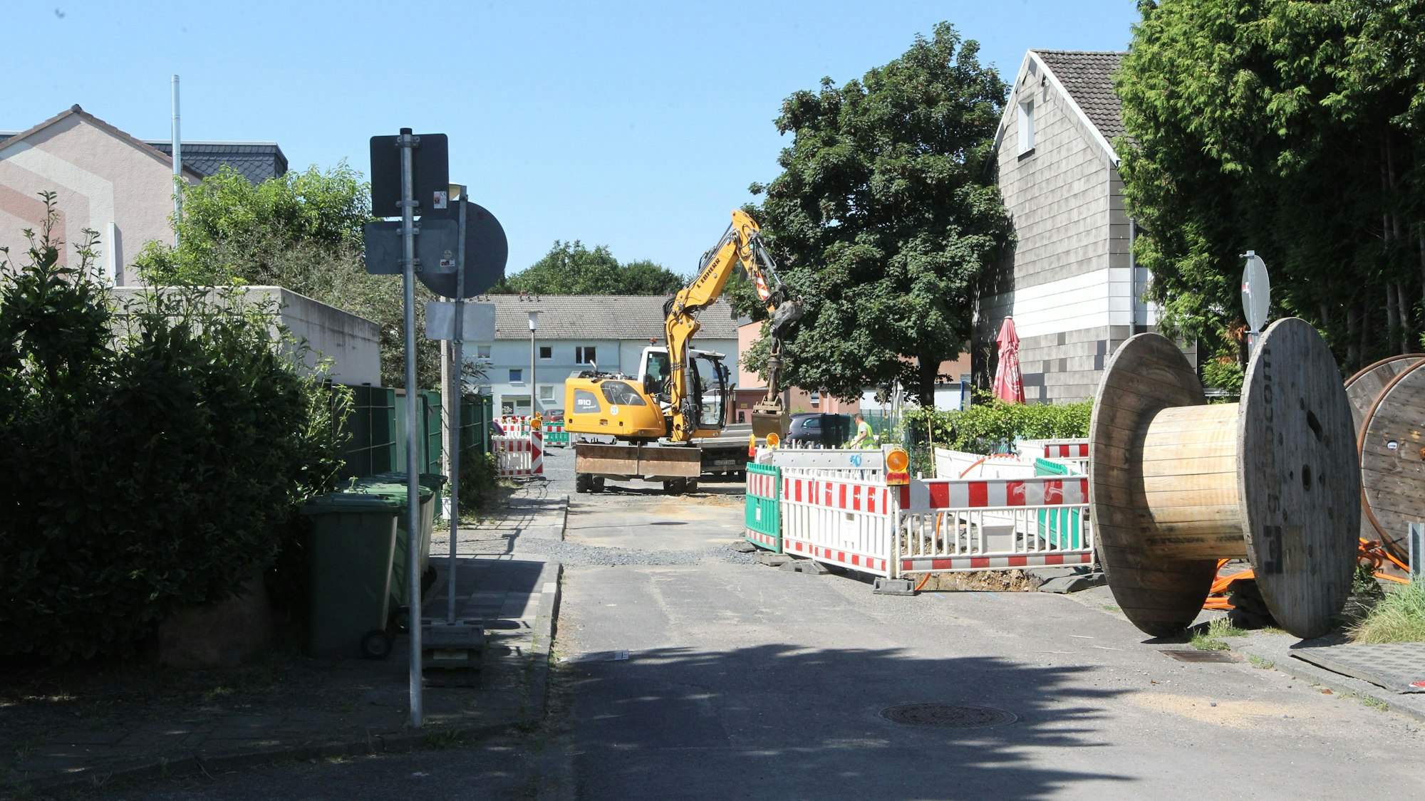 Eine Straßenbaustelle in einem Wohngebiet. Auf der Fahrbahn steht ein gelber Bagger, im Vordergrund liegt eine Kabeltrommel.